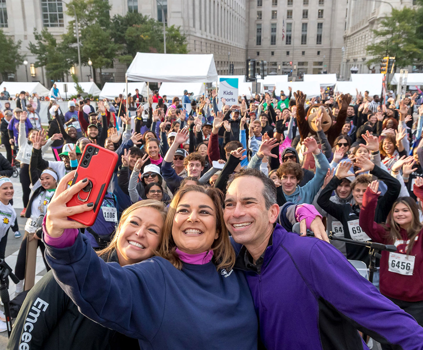 Three people in the foreground take a smiling selfie at an outdoor event, while a large, cheerful crowd behind them waves and poses, surrounded by white tents and city buildings.